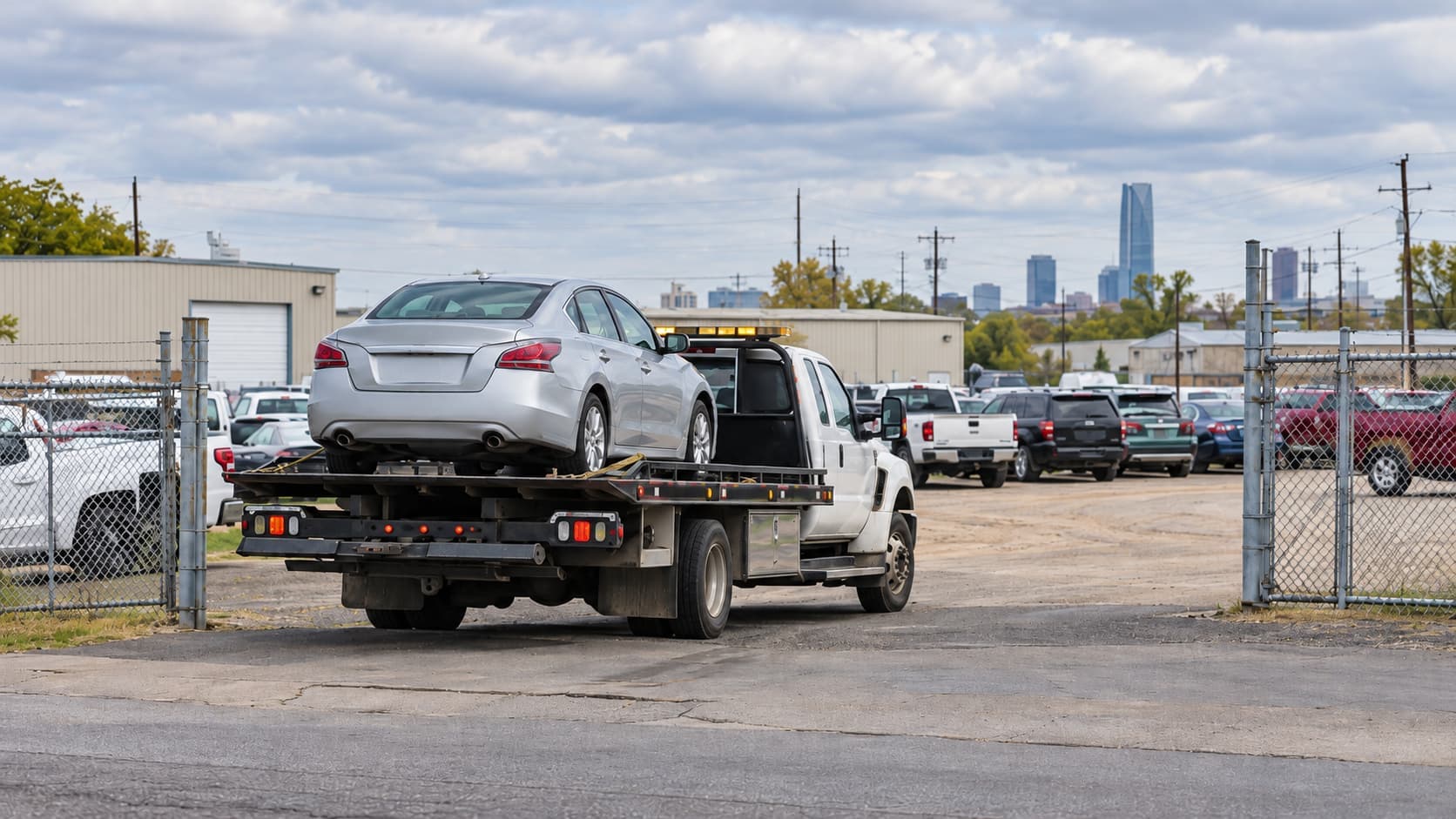 Flatbed tow truck transporting a silver sedan into a fenced vehicle impound lot in Oklahoma City, with parked vehicles and the downtown skyline in the background.